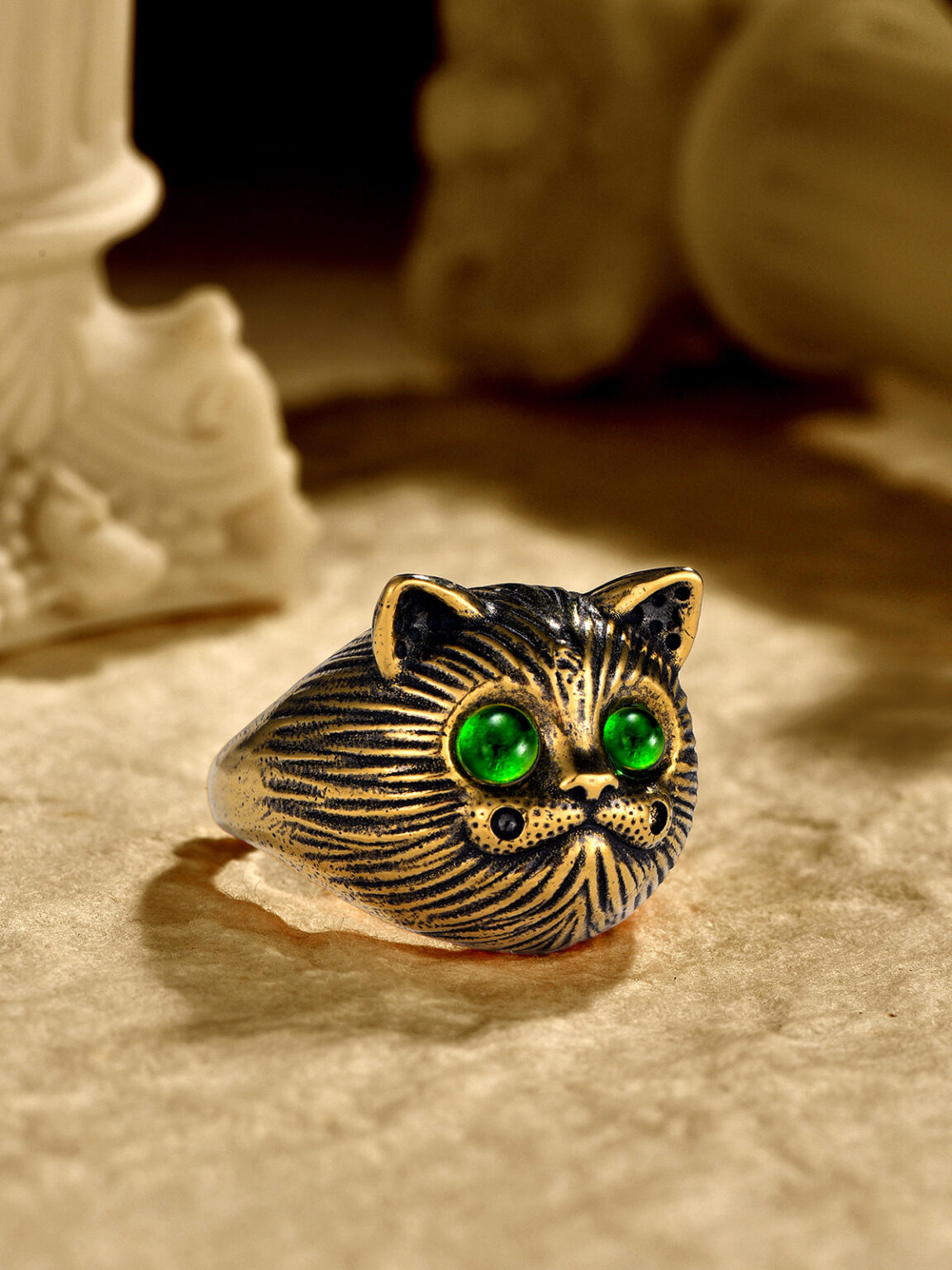 Gold owl ring with green gemstone eyes photographed on a warm textured background.