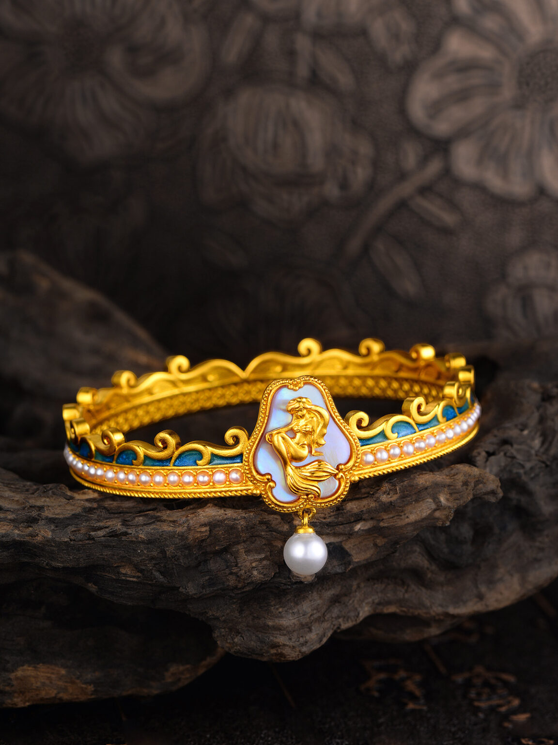 Ornate gold bracelet with enamel details and a pearl accent on a dark patterned background.
