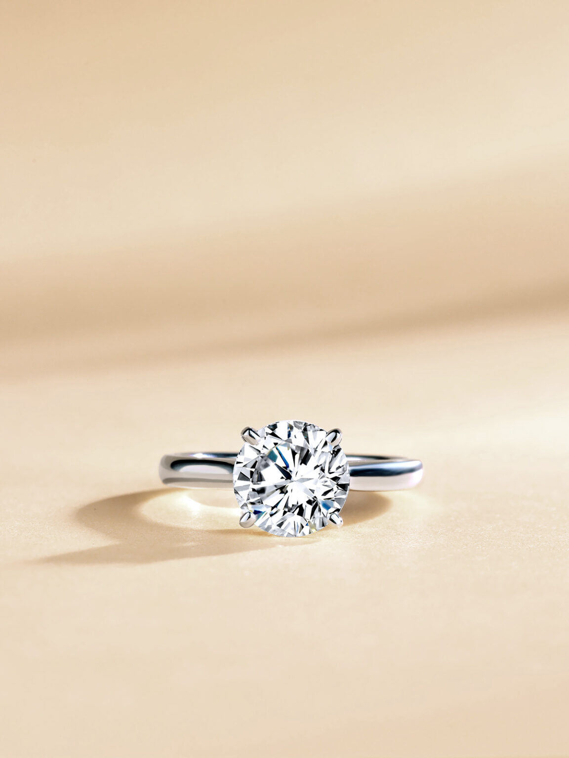 Solitaire diamond engagement ring photographed on a soft champagne beige background.