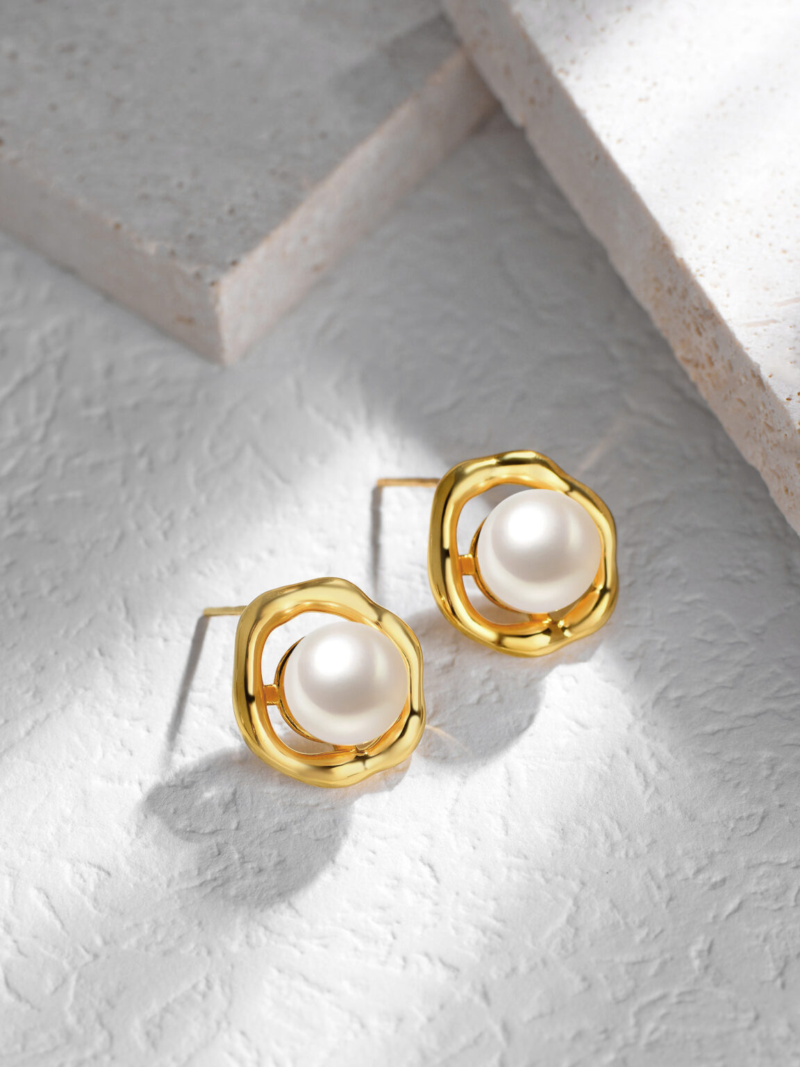 Gold pearl stud earrings displayed on a light gray textured stone surface.