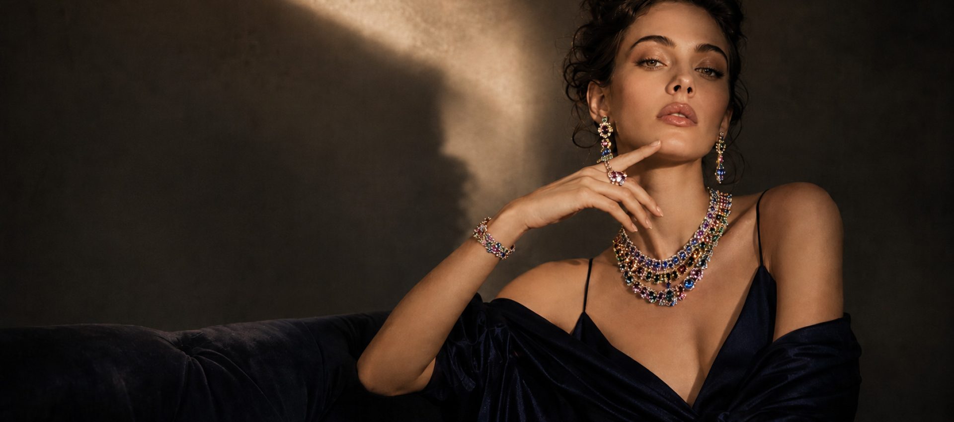 Home Female model in a dark satin gown wearing layered multicolor gemstone necklaces, drop earrings, bracelet and rings against a dramatic shadowed background.