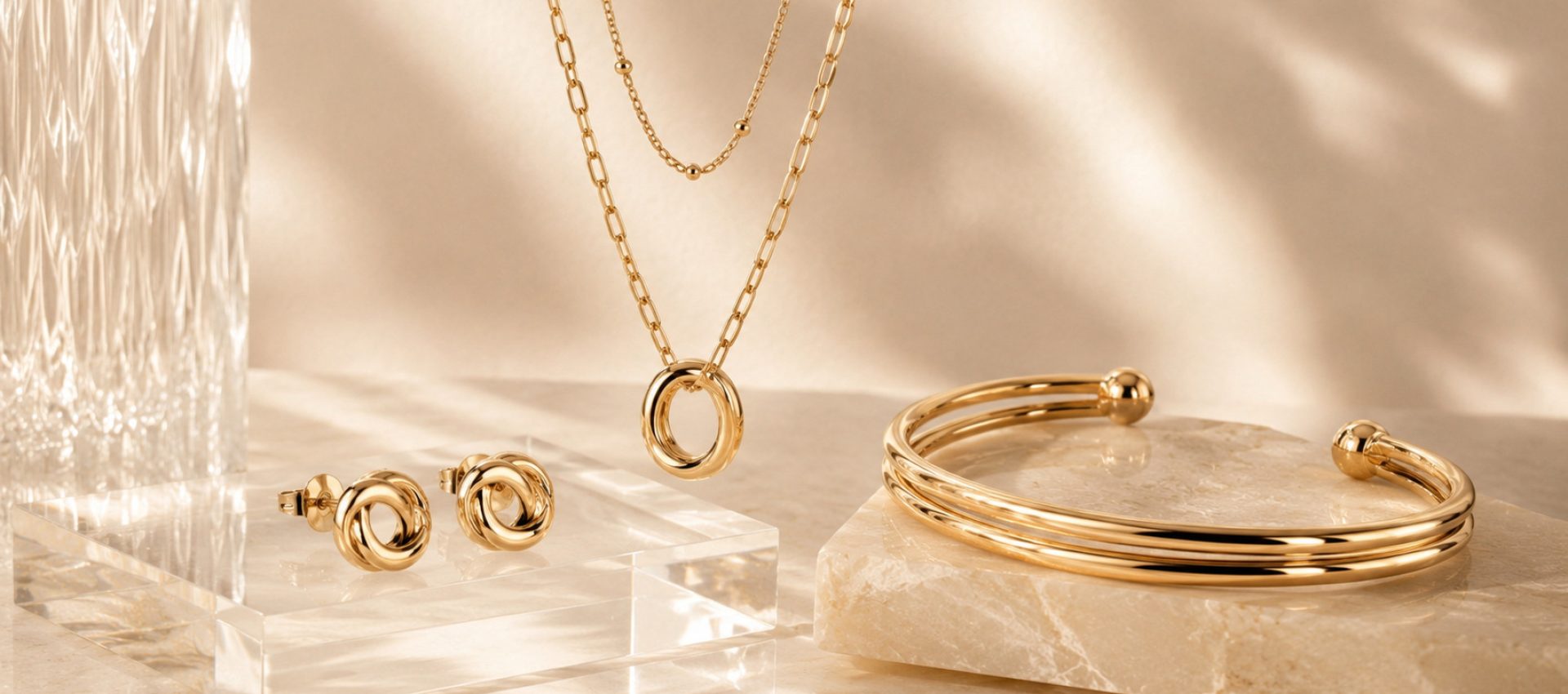 Home Layered gold-tone necklaces, knot stud earrings and an open cuff bracelet displayed on clear acrylic and marble props in warm natural light.